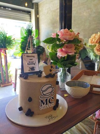 Coffee-themed grand opening cake displayed beside pink flowers inside a café