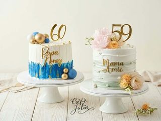 Two white milestone birthday cakes on stands: one with blue brushstrokes and a gold 60 topper, the other with pastel flowers and a gold 59 topper.
