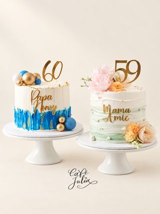 Two white milestone birthday cakes on stands: one with blue brushstrokes and a gold 60 topper, the other with pastel flowers and a gold 59 topper.